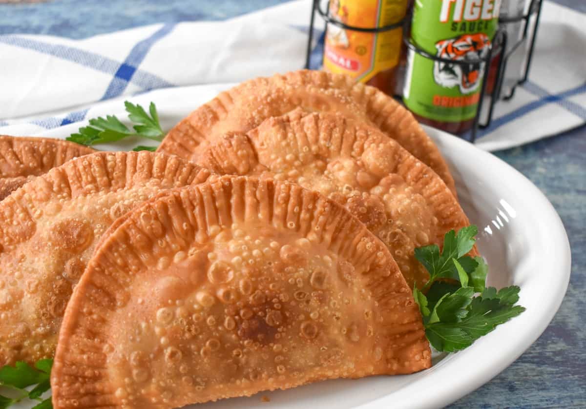 Four turkey empanadas arranged on a white platter and garnished with fresh parsley. In the back ground there are bottles of hot sauce.