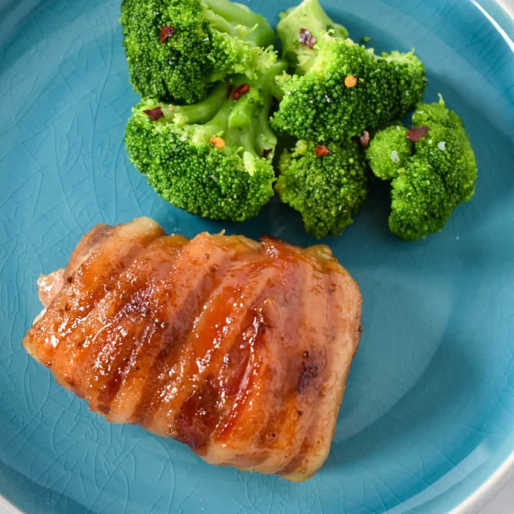 The bacon wrapped chicken thighs served on a aqua colored plate with a side of steamed broccoli garnished with a little crushed red pepper.