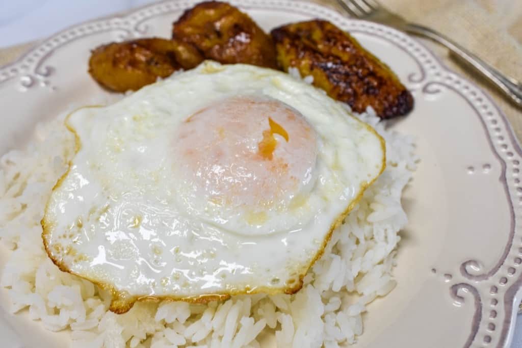 A fried egg on a bed of white rice with fried sweet plantains on the side served on an ornate off-white plate.