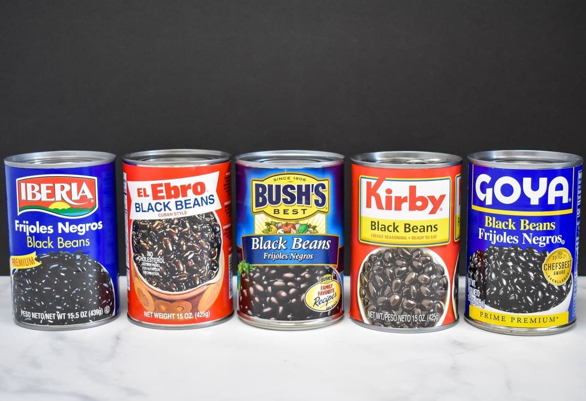 Five cans of black beans all different brands set on a white table with a black background.