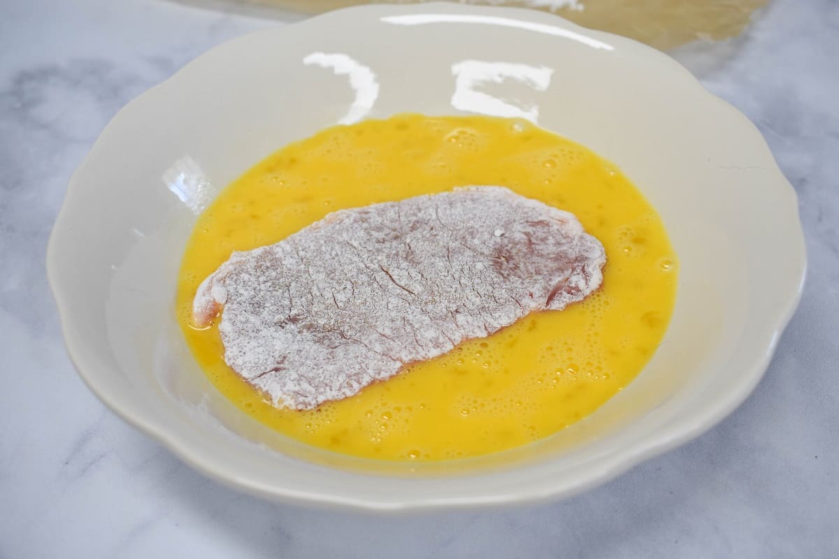 A pork chop that's dredged in flour set in a white bowl with egg.