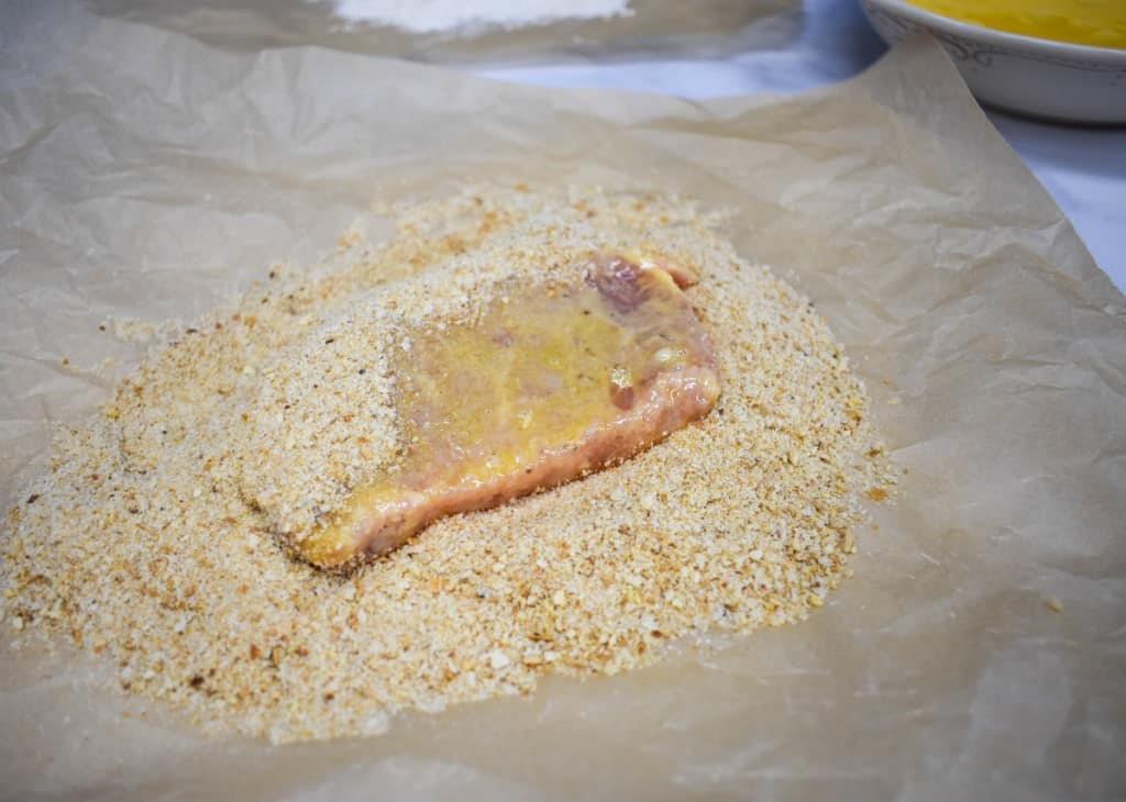 A boneless pork chop being covered in breadcrumbs on a large piece of parchment paper.