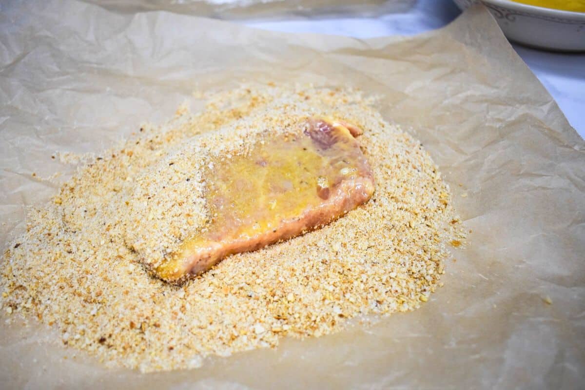 A boneless pork chop being covered in breadcrumbs on a large piece of parchment paper.