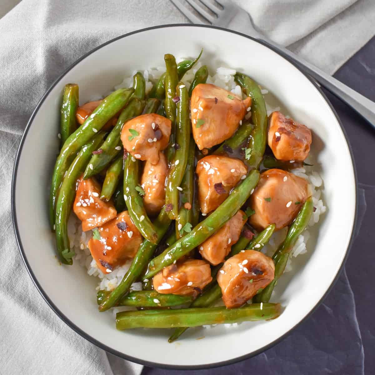 The chicken and green bean stir fry served in a white bowl with a black rim with a beige linen on a black table. There is a fork to the upper right side.