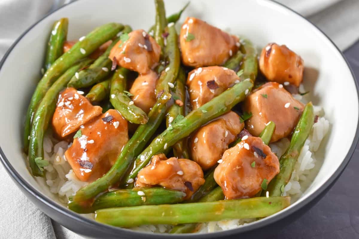 A close up image of the chicken and green beans served on a bed of white rice in a white bowl with a black rim.