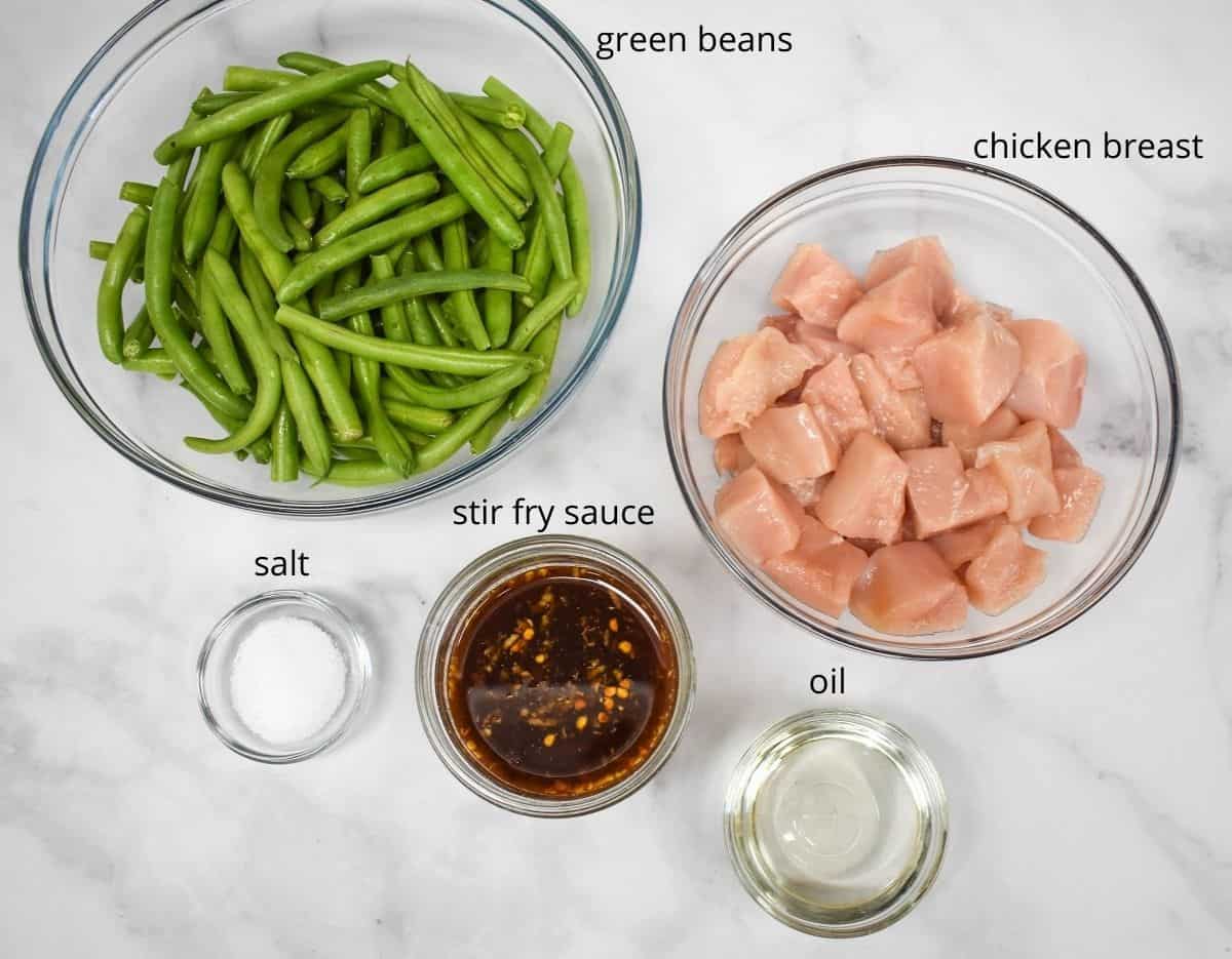 The ingredients for the dish arranged in glass bowls on a white table. The stir fry sauce is already prepared and there is a label next to each ingredient with the name.
