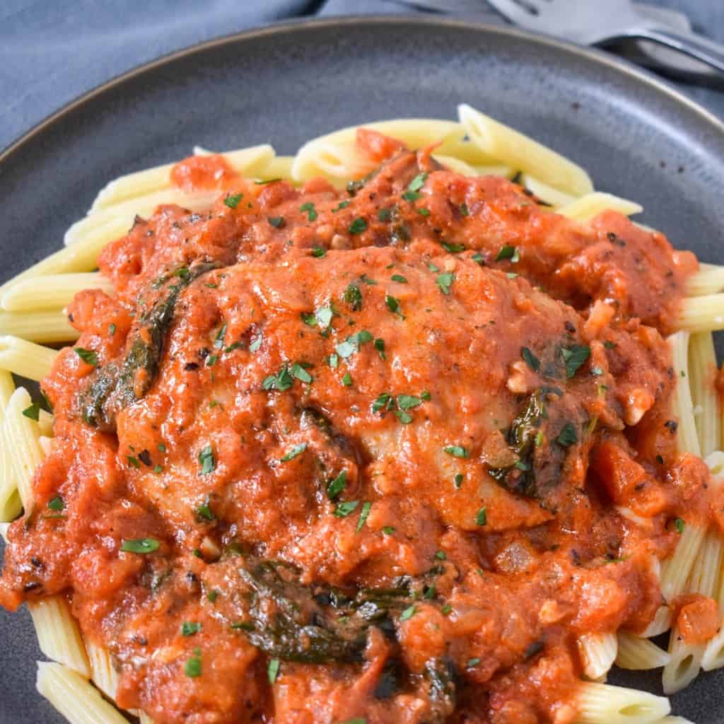 A close up of the finished dish served on a bed of penne pasta on a gray plate and garnished with chopped parsley.