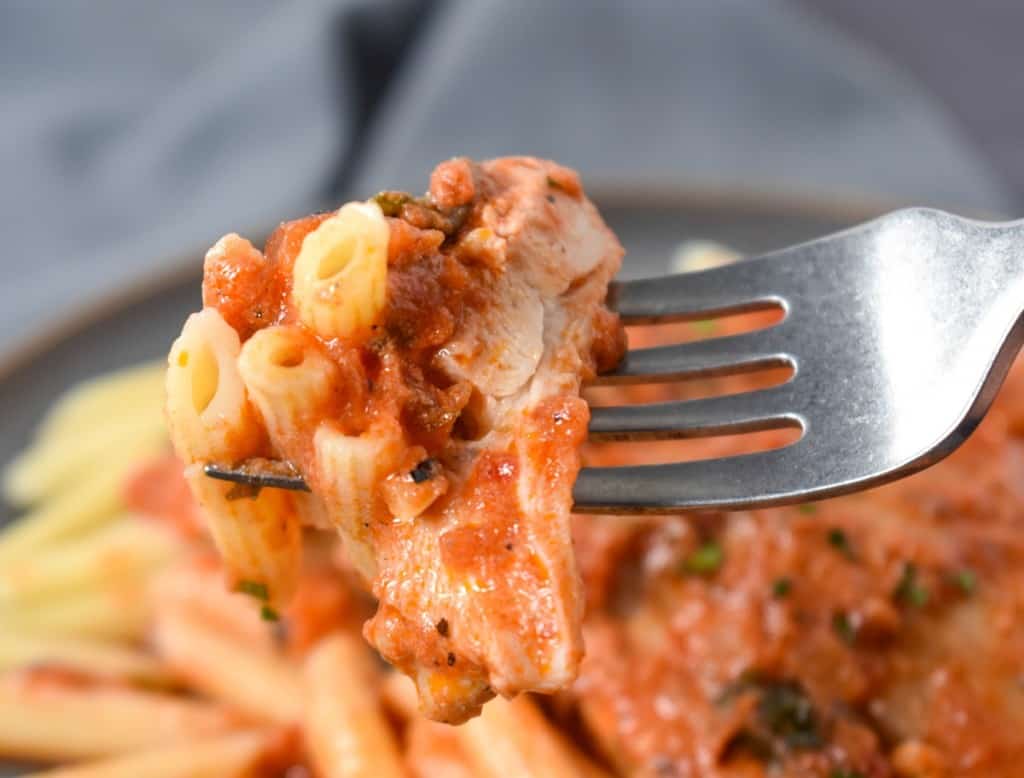 A close up of a slice of chicken and pasta covered in tomato sauce on a fork held over the finished plate.