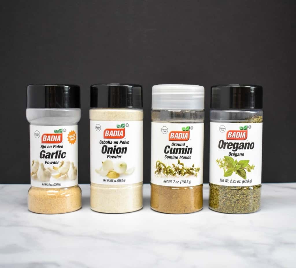 Containers of garlic powder, onion powder, cumin and dried oregano lined up on a white table with a black background.