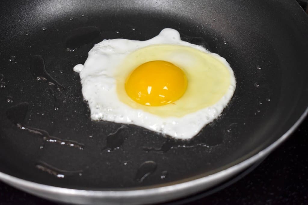 An egg cooking in a non-stick skillet, the yolk is not cooked but the egg white is almost set around it.