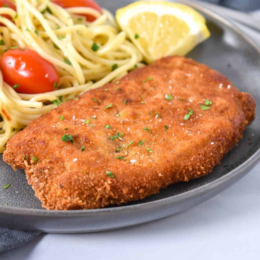 An image of a breaded fried pork chop with spaghetti and small tomatoes in the background and a lemon wedge on the side. The meal is served on a gray plate set on a white table.