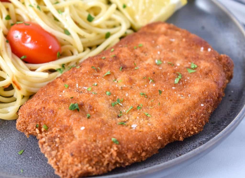 An image of a breaded fried pork chop with spaghetti and small tomatoes in the background and a lemon wedge on the side. The meal is served on a gray plate.