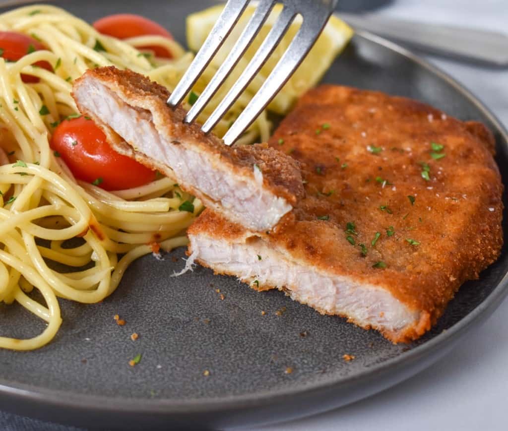 A close up image of the cut piece of fried pork chop held up by a fork. In the background is the remaining piece served with spaghetti with tomatoes. The meal is served on a gray plate.