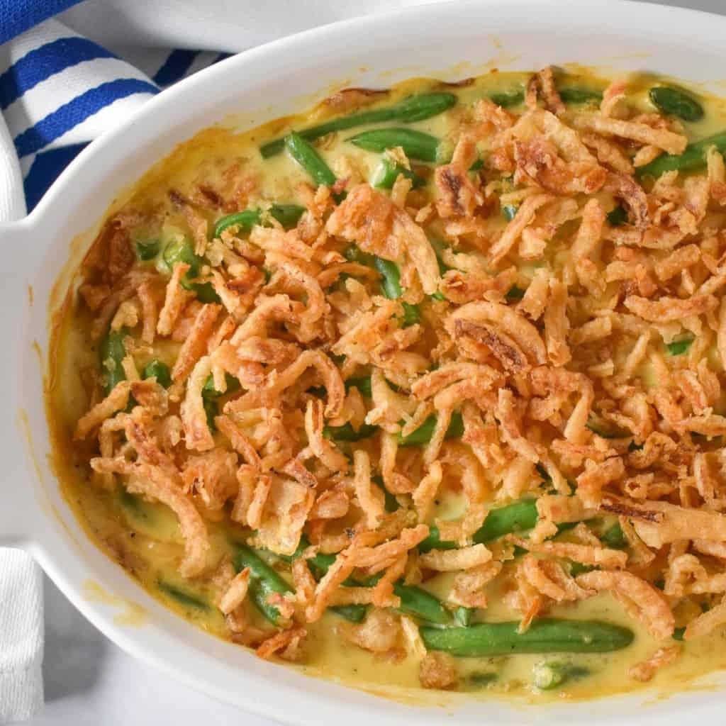 The green bean casserole in a white casserole dish with a blue and white striped kitchen towel in the background.