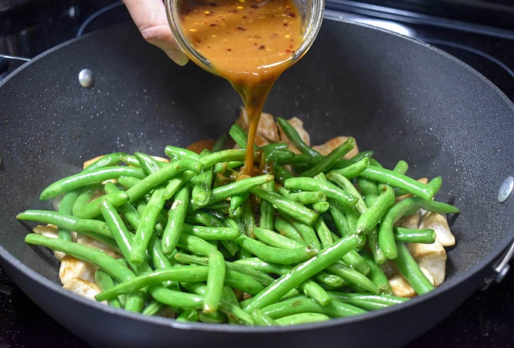 The green beans on top of the chicken in a large, black skillet and the stir fry sauce being added.