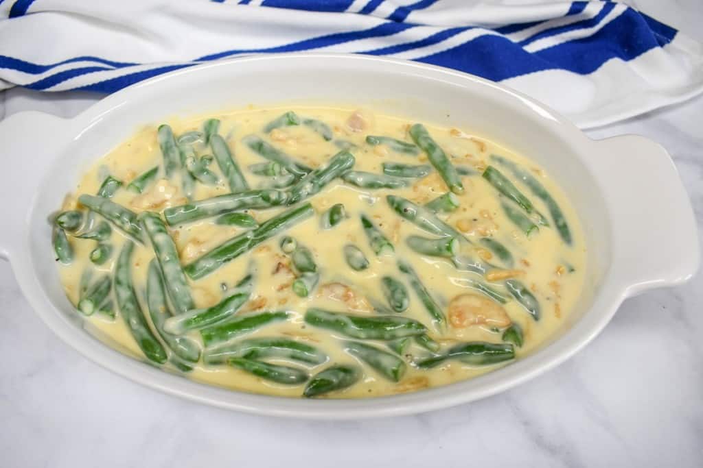 The creamy sauce, green beans and crispy onions after stirring in a white casserole dish. There is a blue and white striped towel in the background.