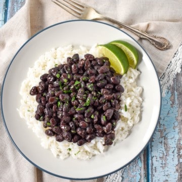 An image of black beans over white rice with two lime wedges, served on a white plate with a blue rim. The plate is set on a blue wood table with a beige linen and a fork to the top right.