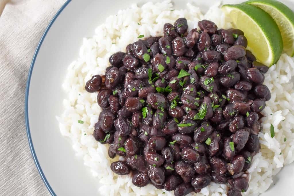 A close-up image of the black beans served over white rice, garnished with chopped parsley with two lime wedges on the side. The food is served on a white plate with a light blue rim.