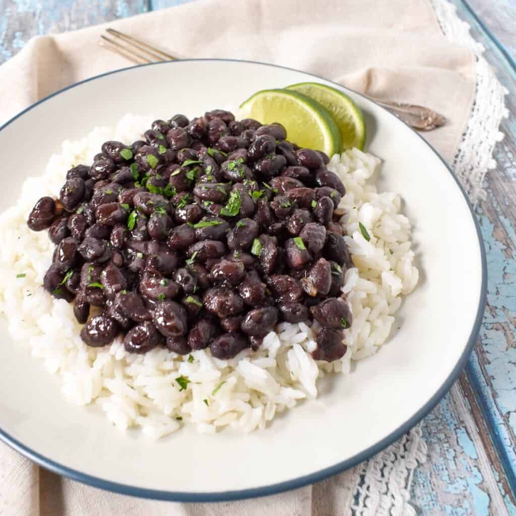 The black beans served over white rice, garnished with chopped parsley with two lime wedges on the side. The food is served on a white plate with a light blue rim.