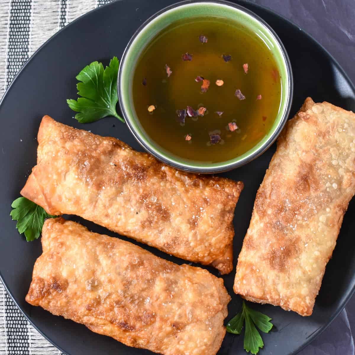 Three egg rolls set on a black plate with a small green bowl with sauce in it.