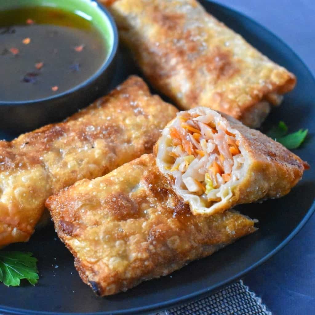 An image of the egg rolls set on a black plate with a green bowl with sauce. One of the egg rolls is cut in half showing the vegetable filling.