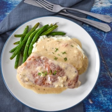 An image of the pork chop served with cream sauce on a bed of mashed potatoes and a side of green beans. The meal is served on a white plate set with a gray linen, fork and knife on a blue table.