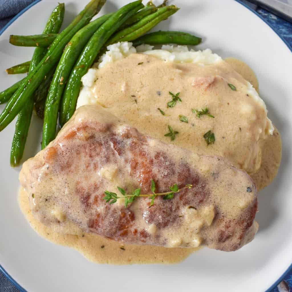 A close up image of the creamy pork chop served on a bed of mashed potatoes with a side of green beans on a white plate.