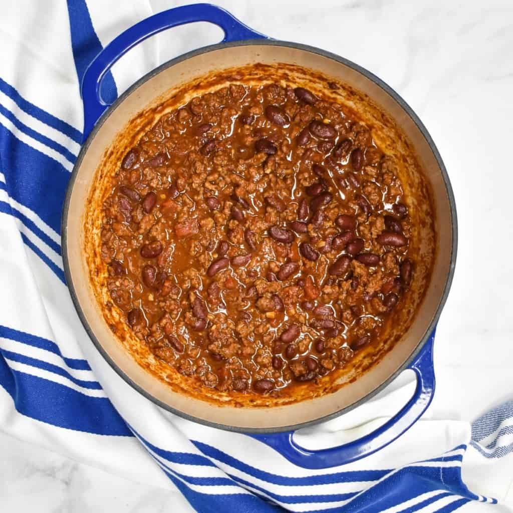 The cooked chili in a blue pot set on a white table with a blue and white striped kitchen towel.