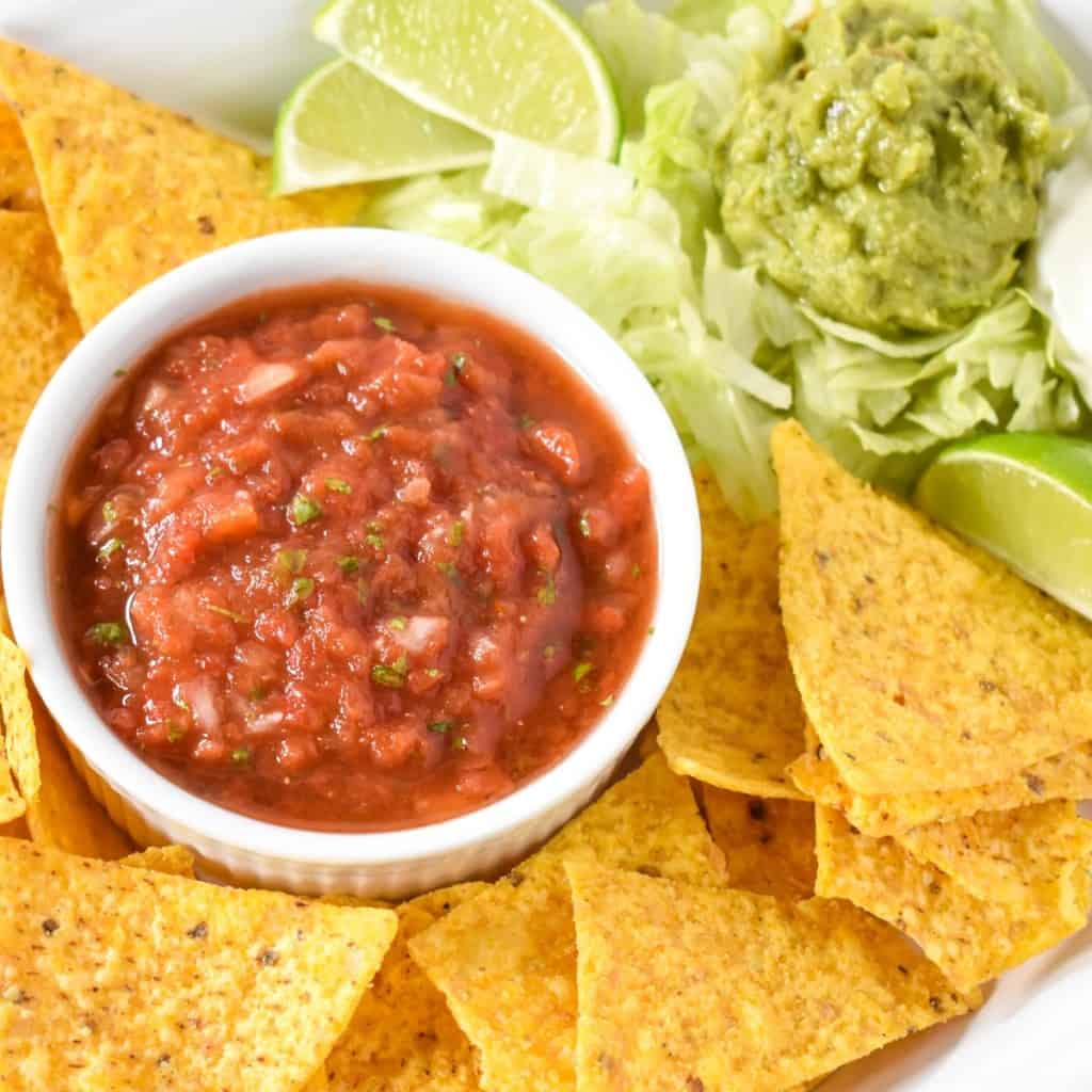 The restaurant style salsa served in a white bowl that's set on a platter with tortilla chips and a bed of lettuce, guacamole and lime wedges on the top, right side.