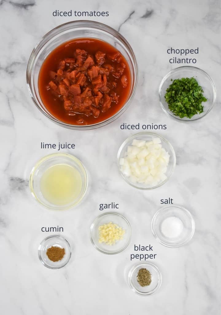 The ingredients for the salsa prepped and arranged in glass bowls on a white table. Each ingredient has a label in black letters above it.