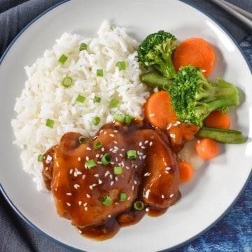 An image of a chicken thigh served with sauce and garnished with sliced green onions and sesame seeds. It is served with white rice and a side of mixed vegetables with broccoli and carrots.