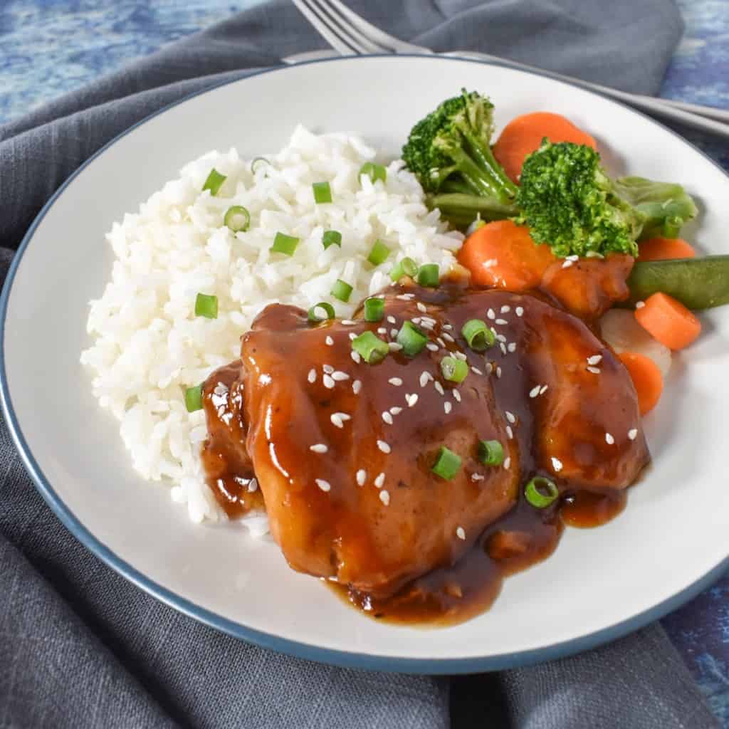 An image of the finished dish served on a white plate with a side of white rice and broccoli and carrots. The chicken and rice are garnished with sliced onions and sesame seeds.