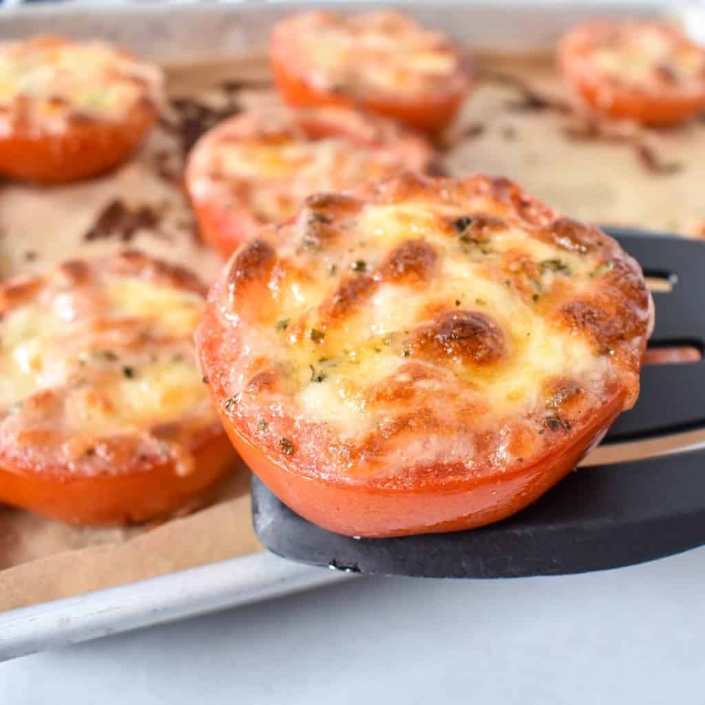 A baked tomato piece held up by a black spatula. The remaining tomatoes can be seen in the background on a parchment paper lined baking sheet.