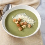 An image of the cream of spinach soup served in a white bowl and garnished with parmesan cheese and croutons. The plate is set on a white table with a beige linen.