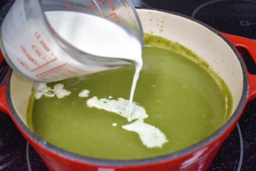 An image of the spinach soup in a red pot with half and half being added with a glass measuring cup.