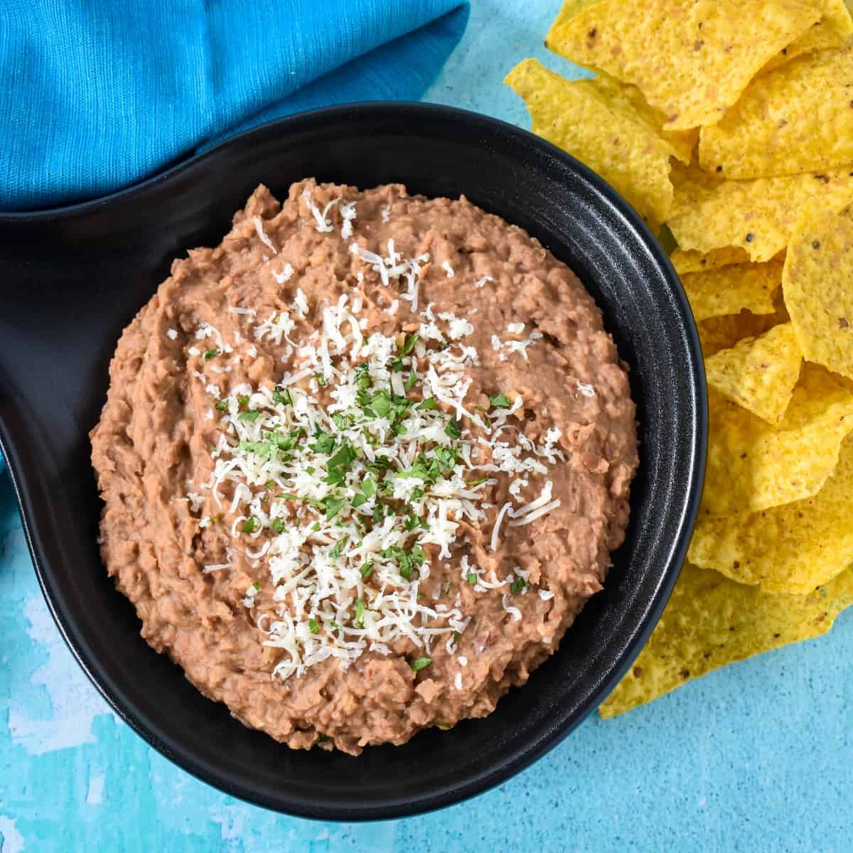 Refried beans served in a black serving skillet with corn tortilla chips to the right and an aqua colored linen set on a light blue board.