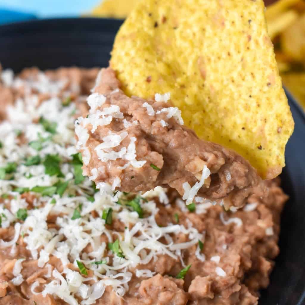 Refried beans garnished with grated cheese and chopped cilantro and being scooped with a corn tortilla chip.