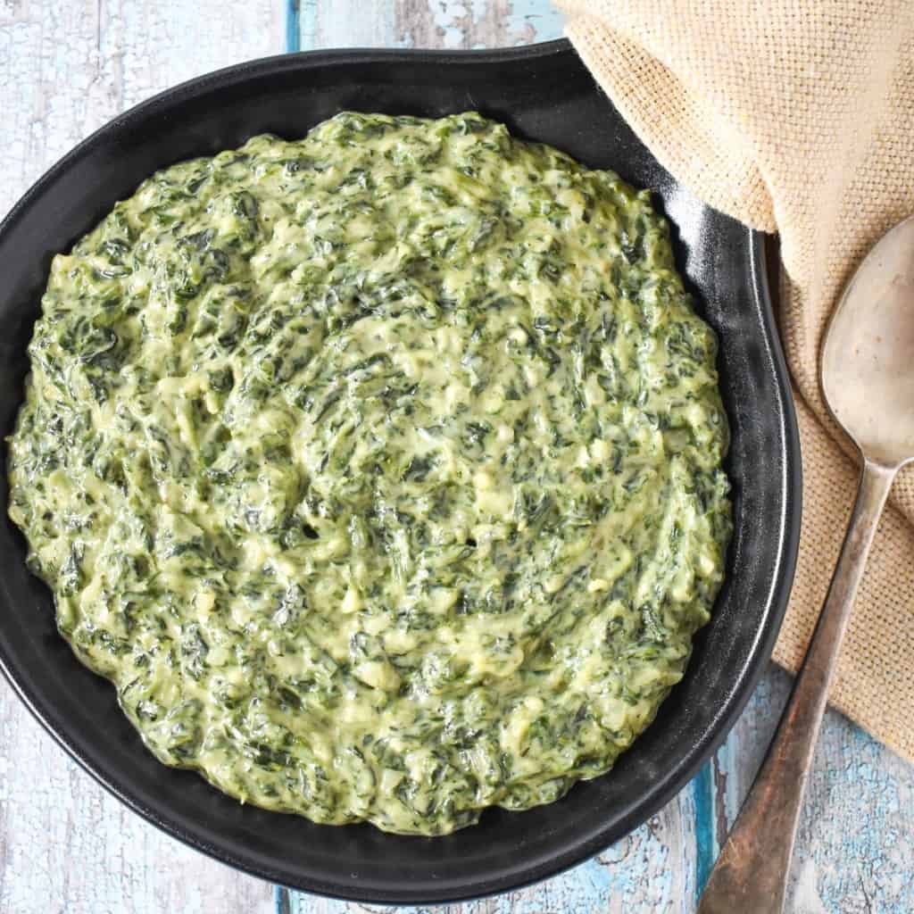 The creamed spinach served in a black serving skillet with a beige linen and serving spoon to the right side.