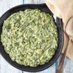 The creamed spinach served in a black serving skillet with a beige linen and serving spoon to the right side.