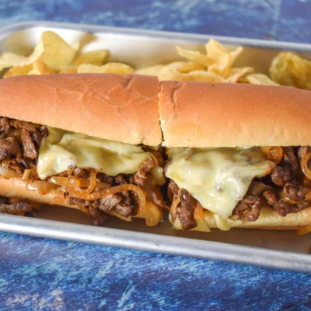 An image of the steak sandwich served on a small sheet pan with potato chips and set on a blue table.