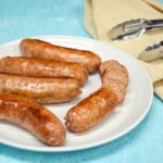 An image of five links of Italian sausage served on a white plate set on a light blue table with a beige kitchen towel and tongs on the right side.