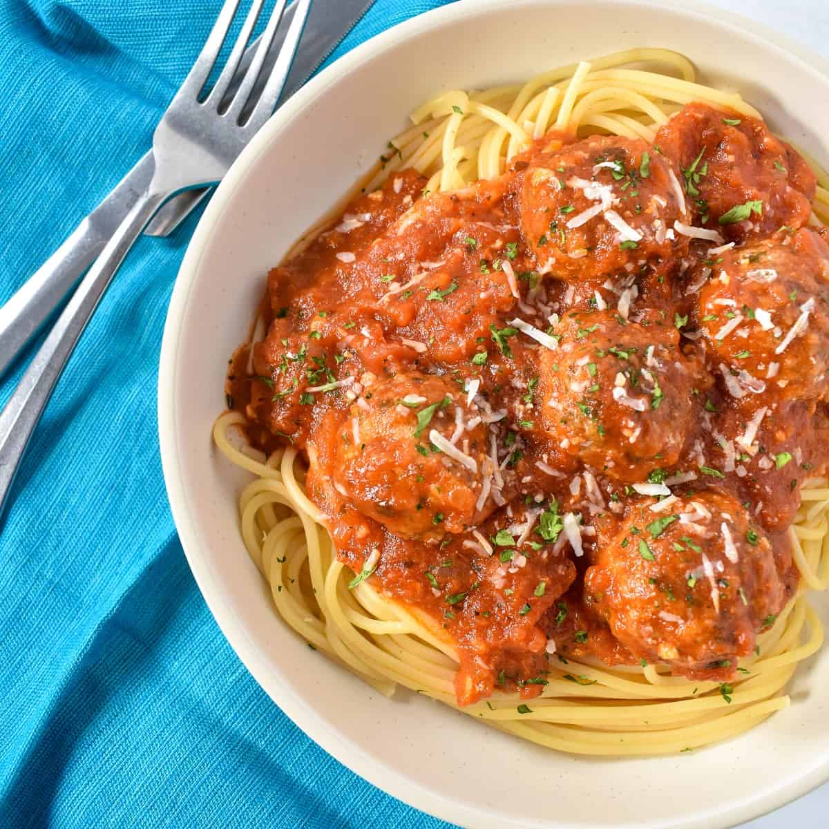 The meatballs served on a bed of pasta in a white bowl and set on an aqua colored linen.