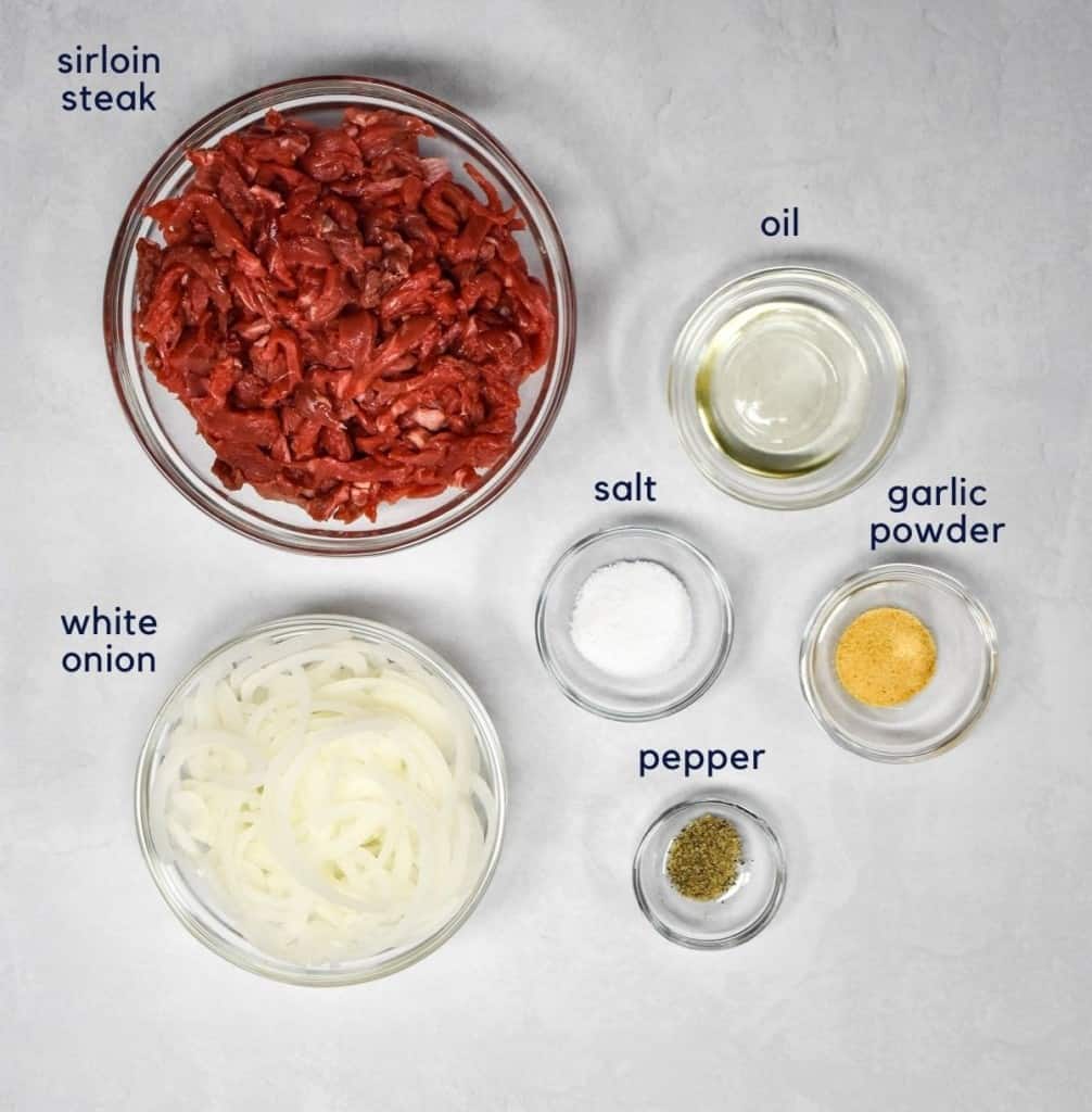 The ingredients for the steak and onions arranged in glass bowls set on a white table. Each ingredient has a small label with its name on the top of side.