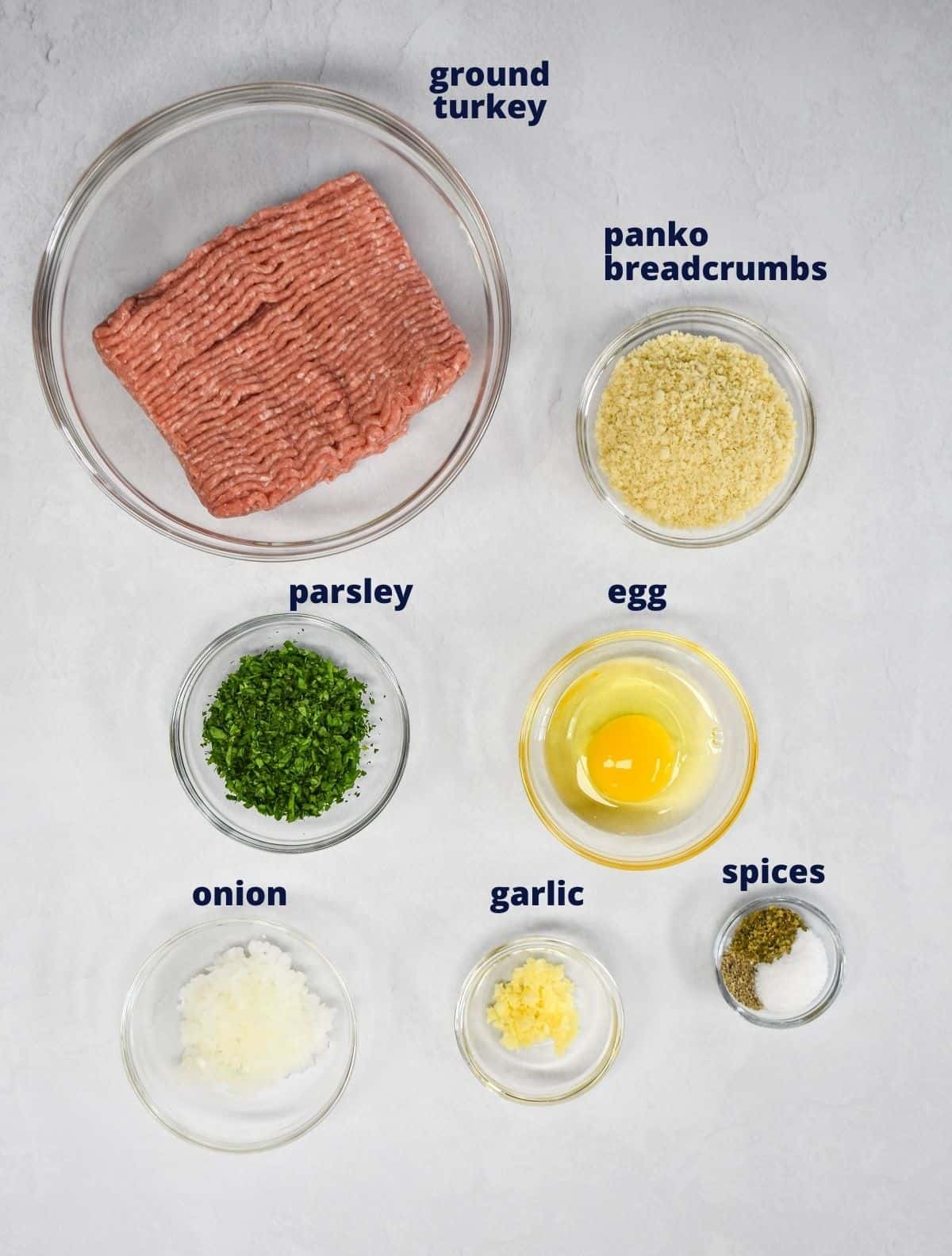 The ingredients for the meatballs prepped and arranged in glass bowls on a white table.