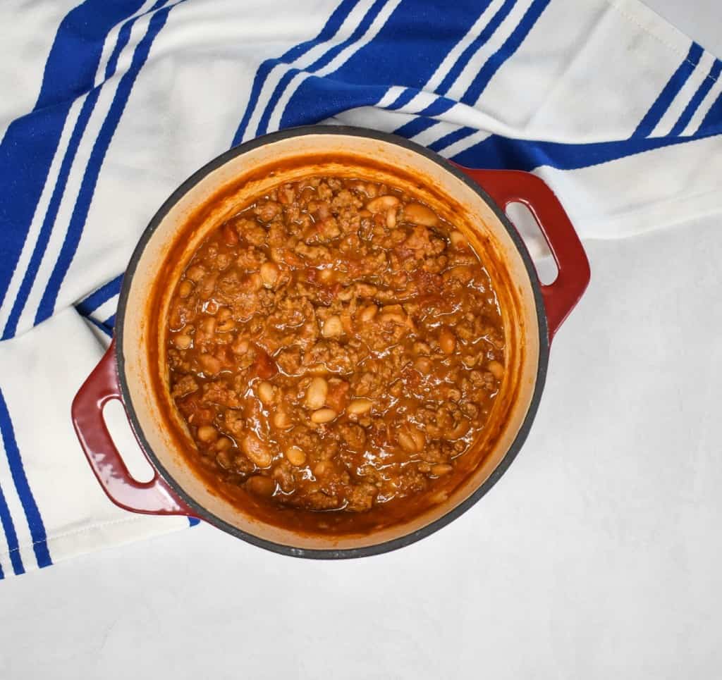 An image of the turkey white bean chili in a red pot after it is cooked and reduced. The pot is set on a white table with a blue and white striped kitchen towel.