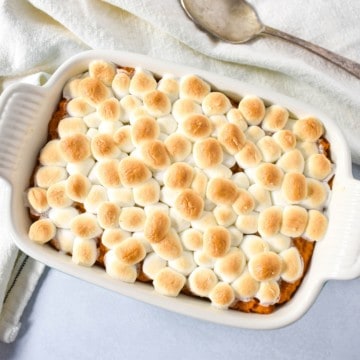 An image of the sweet potato casserole in a white dish set on a white table with a beige linen and serving spoon.