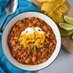 An image of the turkey white bean chili served in a white bowl with a black rim. Alongside the chili is an aqua linen, a spoon and a small cutting board with tortilla chips and lime wedges.