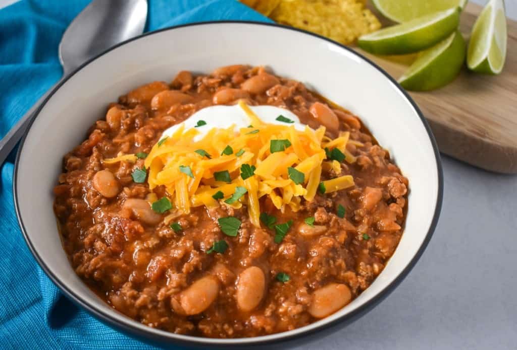 A close up image of the chili garnished with shredded cheese, sour cream and chopped parsley. It is served in a white bowl with a black rim. Alongside the chili is an aqua linen, a spoon and a small cutting board with tortilla chips and lime wedges.