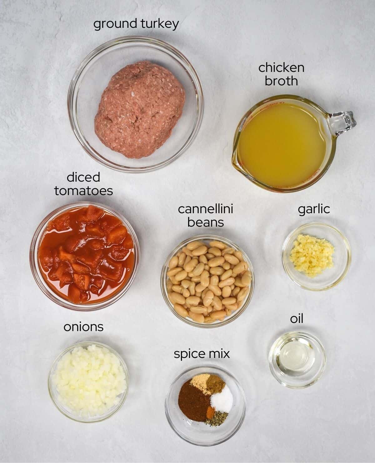 An image of the ingredients for the chili arranged in glass bowls and set on a white table. Each ingredients has a small label with the name in black letters.