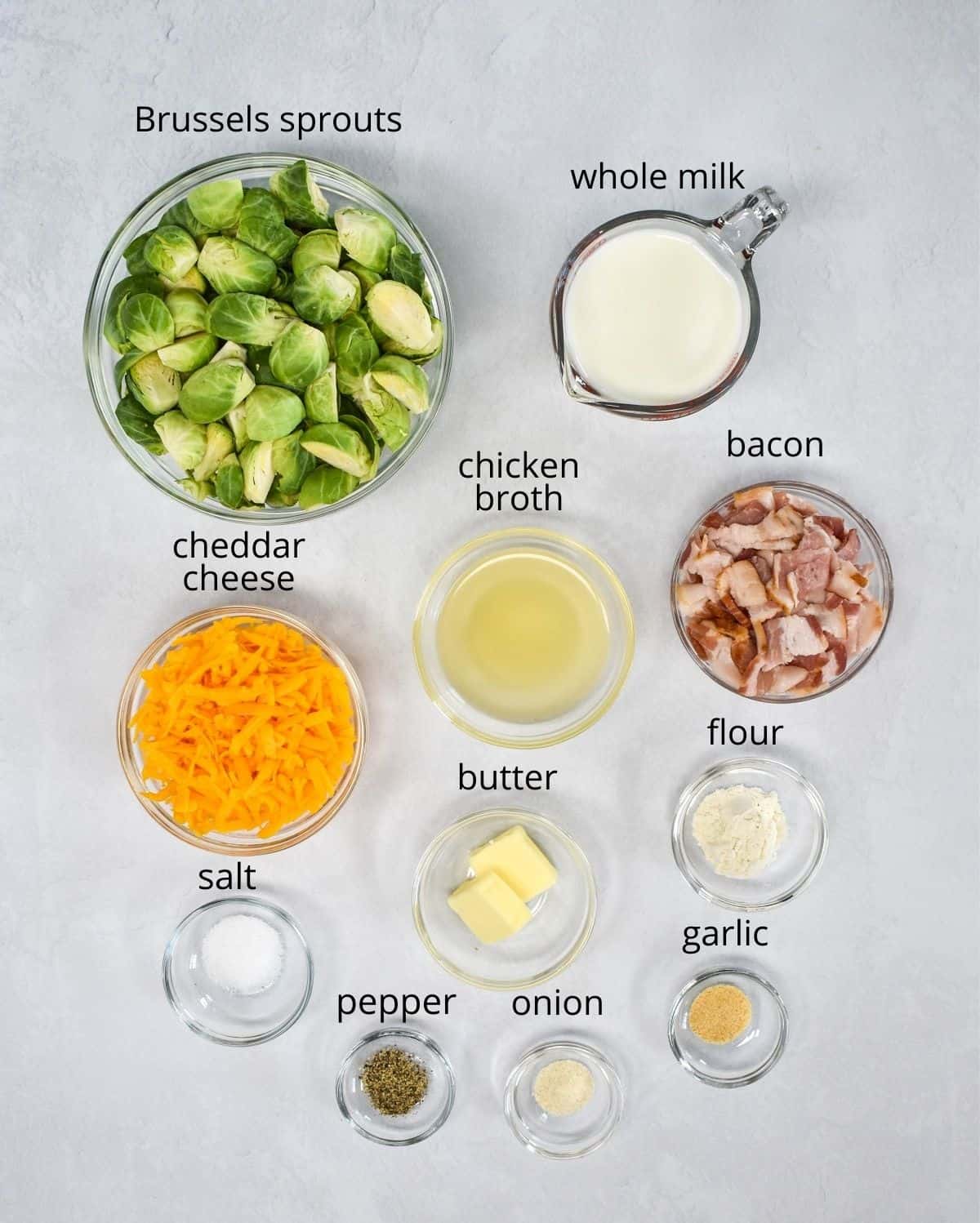 The ingredients for the casserole prepped and arranged in glass bowls and set on a white table. Each ingredient has a small label with the name above it in small black letters.
